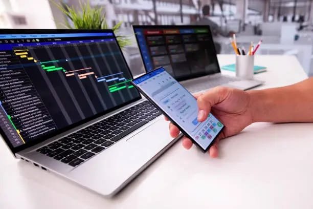 "Person using a smartphone for financial analysis with data displayed on two laptops"