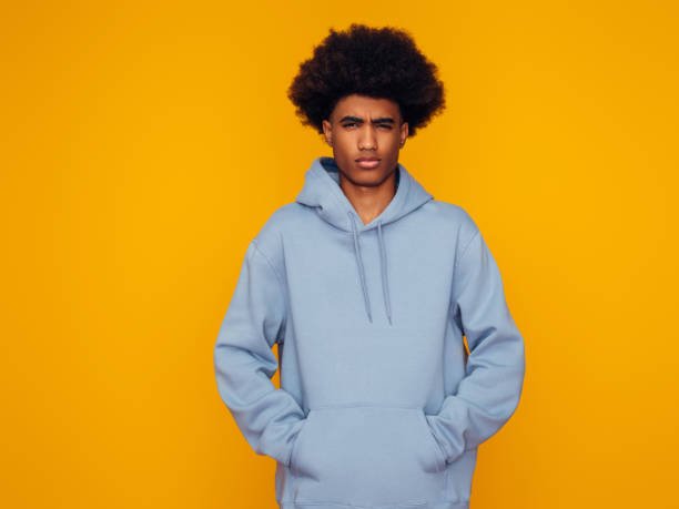 Young person wearing a light blue hoodie with an afro hairstyle against a bright yellow background.