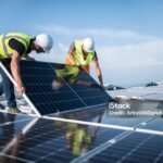 "Two workers installing solar panels on a rooftop under clear skies"