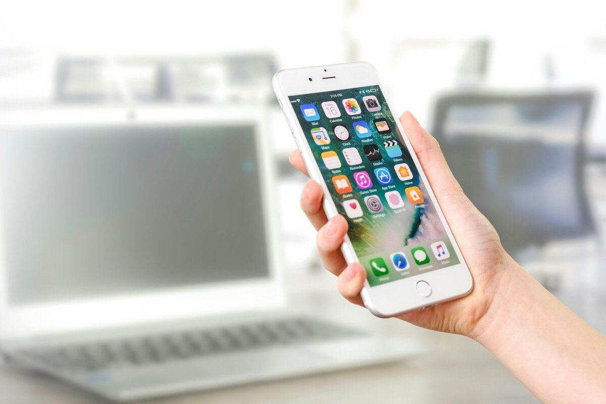 "Hand holding a smartphone with app icons displayed in front of a blurred laptop"