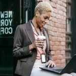 "Professional woman holding coffee while working on a laptop outside a coffee shop"