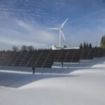 Solar panels in a snowy field with a wind turbine in the background.