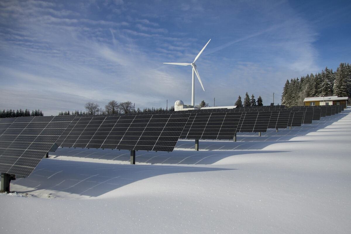 Solar panels in a snowy field with a wind turbine in the background.