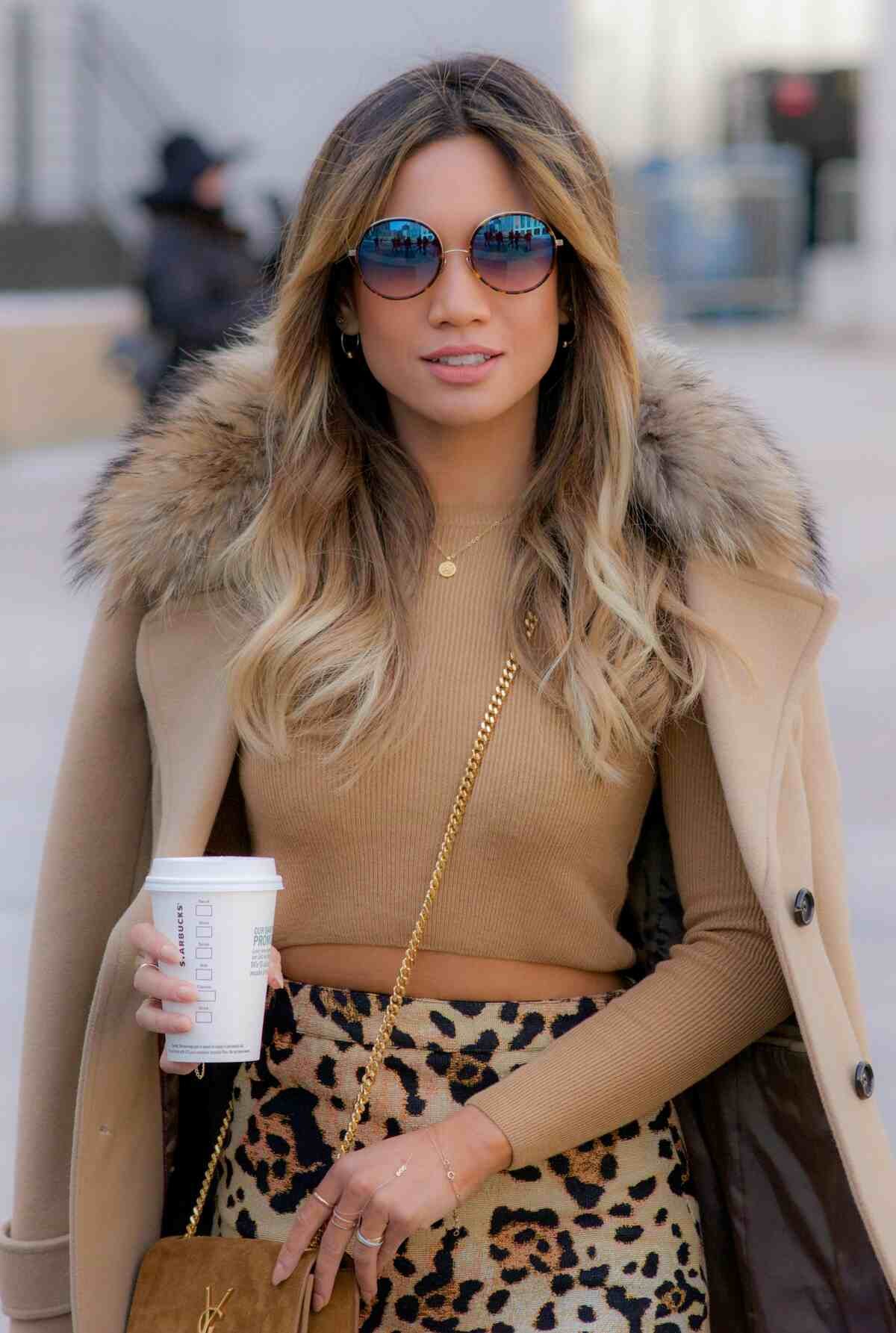 Fashionable woman in a beige coat with fur collar, holding coffee and wearing sunglasses"