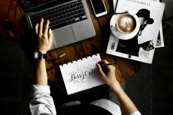 Overhead view of a person working at a wooden table with a laptop, coffee, and magazines, hand-drawing a design.