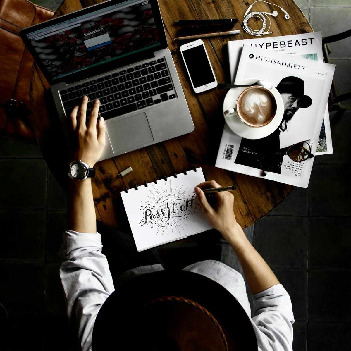 Overhead view of a person working at a wooden table with a laptop, coffee, and magazines, hand-drawing a design.