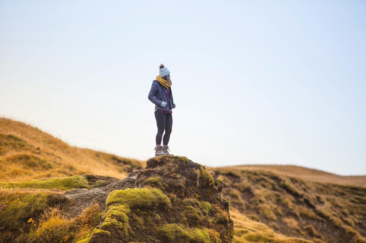 "Person standing on a moss-covered rock in a wide-open grassy landscape, wearing a winter hat, scarf, and jacket, looking into the distance"