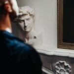 A man looking into a mirror with a classical bust statue in the background, evoking themes of reflection and self-awareness.