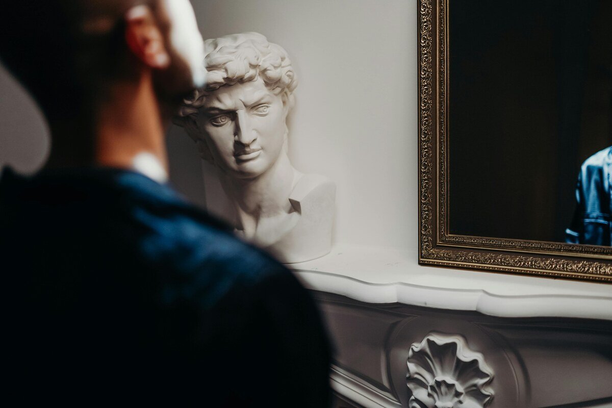 A man looking into a mirror with a classical bust statue in the background, evoking themes of reflection and self-awareness.