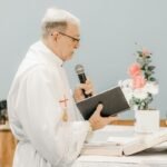 Elderly priest delivering a sermon while holding a book and microphone at the altar.