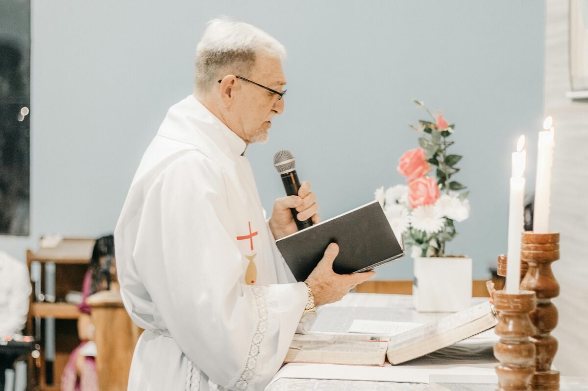 Elderly priest delivering a sermon while holding a book and microphone at the altar.